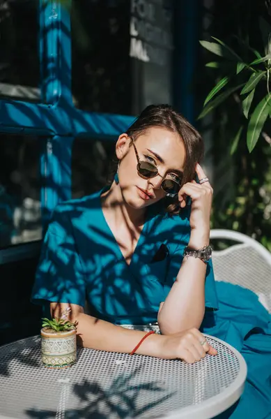 stock image Young woman in sunglasses relaxes at an outdoor cafe with a small potted plant, surrounded by shadows and greenery.