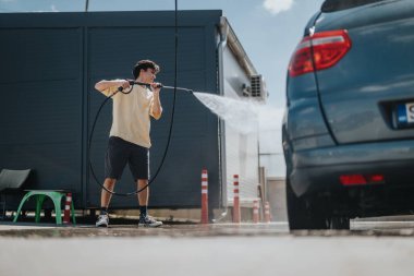 A young man uses a high-pressure water hose to clean a car at a self-service car wash. The setting is outdoors on a sunny day, showcasing diligence and vehicle maintenance. clipart