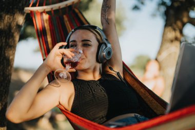 Woman enjoying summer in nature, relaxing on a hammock with a glass of wine and listening to music. Outdoor leisure and relaxation concept. clipart