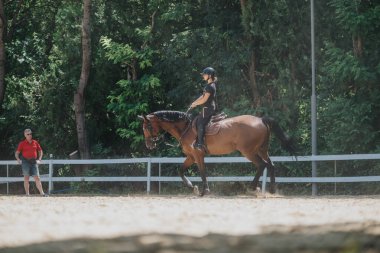 Woman training horse under the supervision of a trainer in an outdoor equestrian arena surrounded by nature. clipart