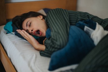 A woman wearing striped pajamas yawns while lying in bed. The scene conveys a sense of coziness and morning relaxation, emphasizing the comfort and warmth of a peaceful sleep environment. clipart