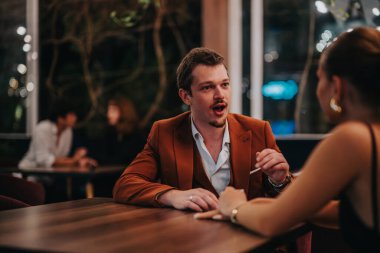 A young couple enjoys a lively conversation at a modern restaurant. The dim lighting and elegant decor create an intimate and engaging atmosphere. clipart