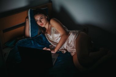 Young woman lying in bed, gazing at her laptop screen at night. The dim lighting creates an intimate and reflective mood, highlighting a moment of solitude and contemplation. clipart