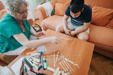 A young boy enjoys a pick-up sticks game with his grandmother in a cozy living room, fostering connection and joy. clipart