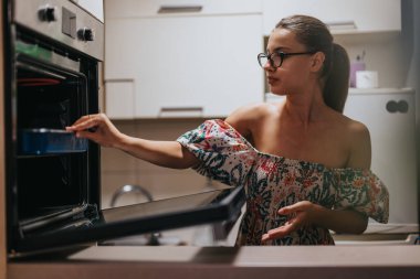 A woman in a colorful dress is baking in a modern kitchen. She is focused as she adjusts the oven, highlighting a home-cooking atmosphere. clipart