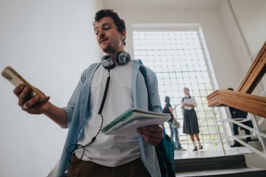 A young student with headphones around his neck checks his smart phone while holding notebooks in a school hallway, with other students in the background. clipart