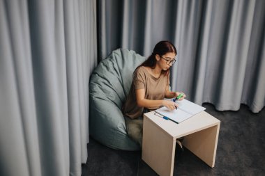 A focused high school student studies in a cozy setting, seated on a bean bag with notebooks and pens, exuding concentration in a tranquil environment. clipart