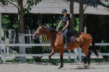 A skilled rider wearing a helmet confidently rides a brown horse in a sunlit outdoor arena, displaying admirable equestrian skills and training in a serene environment. clipart