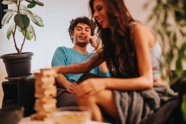 A young man and woman share a laugh while playing a block tower game at home. The warm setting hints at friendship and joy in a relaxed environment. clipart