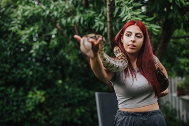 A confident girl with red hair holding a large snake outdoors in a lush garden setting. The image captures a moment of connection and fascination with nature. clipart