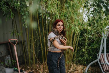 A smiling girl with red hair holding a large snake in a lush garden setting. The scene conveys a sense of adventure and harmony with nature among bamboo plants. clipart