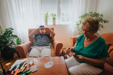 A grandmother and her grandson enjoy a card game in a bright, cozy living room surrounded by plants and comfortable seating. clipart