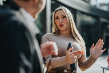 A woman with long blonde hair gestures expressively during a conversation outside. The setting is bright and sunny, capturing a lively interaction between two people in a casual environment. clipart