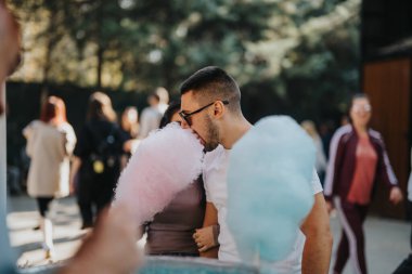 A young man savors pink and blue cotton candy at an outdoor fair. The scene captures a festive atmosphere with blurred people in the background. clipart