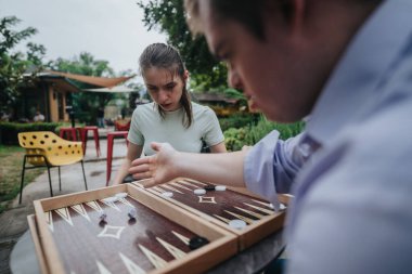 A boy with Down syndrome and a girl in a wheelchair share a playful moment playing backgammon together in a garden setting, promoting inclusivity and friendship. clipart