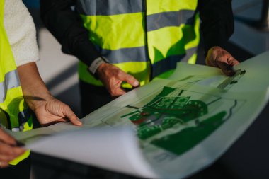 Two architects in high-visibility vests review and discuss building plans at a construction site, highlighting teamwork and construction planning. clipart