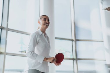 A cheerful businesswoman in a modern office taking a break, enjoying a game of table tennis. The sunlight streaming in enhances her joyful expression and adds energy to the scene. clipart