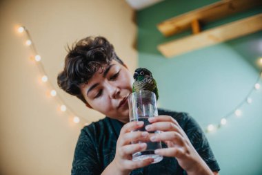 A young girl gently interacts with her parrot perched on a glass, capturing a joyful and heartwarming indoor moment. The scene is lit with warm lights, creating a cozy atmosphere. clipart