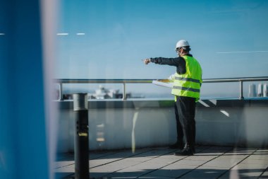 Two architects wearing safety gear discuss building plans while pointing at a construction site. The rooftop view emphasizes city planning and development. The scene conveys professionalism and clipart