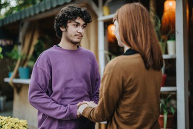 Young couple standing outdoors, engaged in a heartfelt conversation. They are holding hands and looking at each other, conveying trust and connection in a natural setting with plants around. clipart