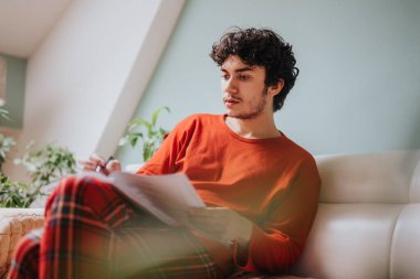 A young man in a red shirt relaxes on a sofa while reading a book. The cozy environment and his plaid pants create a warm and inviting atmosphere. clipart