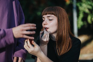 A young woman applies lipstick while using a mirror held by a friend outdoors. The image captures an intimate and casual beauty moment shared between friends in natural light. clipart