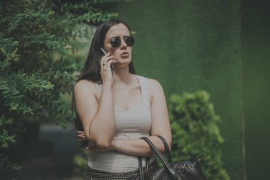 A confident woman stands outside surrounded by greenery, engaged in a phone conversation. She is holding a handbag and wearing sunglasses, exuding professionalism and focus. clipart