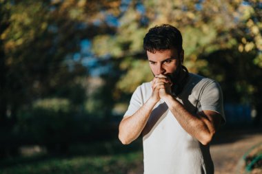 A man stands outside, deep in thought, with sunlight filtering through the trees. His pensive expression highlights a moment of reflection and introspection. clipart