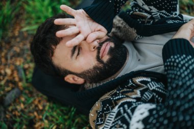 A bearded man wearing a patterned sweater rests on the grass, covering his eyes with one hand. Leaves and greenery surround him, creating a serene and relaxed outdoor setting. clipart