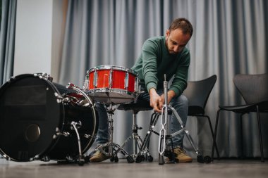 A man prepares a drum kit in a cozy rehearsal room, adjusting the stands and ensuring everything is in place. The black and red drums contrast with the muted background. clipart