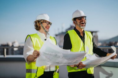 Two architects wearing hard hats and reflective vests study blueprints while laughing on a rooftop. They embody teamwork, planning, and a positive work environment in construction. The cityscape clipart