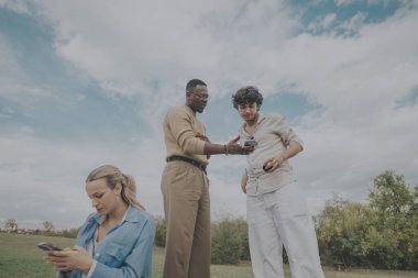 Three young adults engage with their smart phones outside on a sunny day, surrounded by greenery. They appear relaxed and focused, enjoying a moment of technology-driven connection. clipart