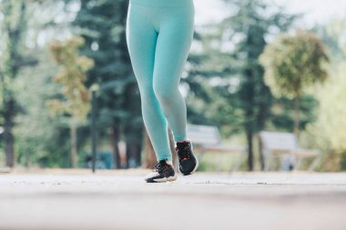 A woman jogs outdoors in a park, enjoying fitness and nature. Her turquoise leggings and black sneakers highlight an active and healthy lifestyle on a sunny day. clipart