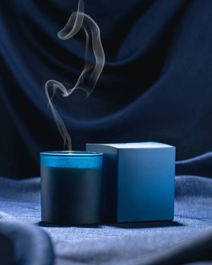 Mavi kutulu mavi kokulu bir mum koyu mavi bir kumaş zeminine karşı tüttürür.