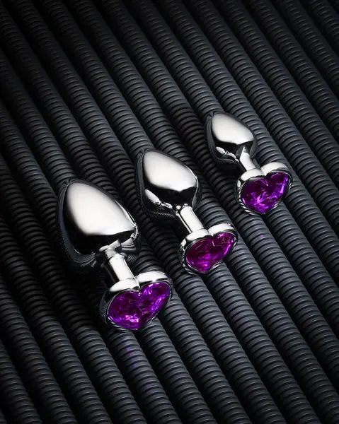 设置三个不同尺寸的银制肛门插头 装饰有心形紫色宝石 镶嵌在有肋的深色背景上 成人性体验性玩具的概念 — 图库照片