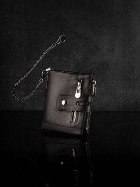 Birden fazla fermuarlı bölmeli bronz deri cüzdan ve dayanıklı dikişlerle süslenmiş hırsızlık önleyici zincir koyu renk yansıtıcı yüzeyde sunuluyor. Şık el yapımı aksesuar