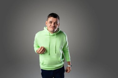 Yeşil kapüşonlu genç bir adam gri bir arka plana karşı duruyor. Yüzünde ciddi bir ifade var ve yumruğunu sıkıyor..