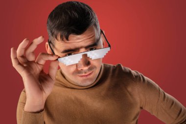 Pikselleştirilmiş güneş gözlüğü takan bir adam kameraya bakar, kahverengi bir kazak giyer ve sağ eliyle gözlüklerini ayarlar. açık kırmızı bir arkaplan 