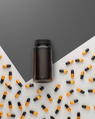 Kapalı, etiketlenmemiş bir şişe beyaz ve gri yüzeyin ortasında duruyor. Dağınık altın ve siyah kapsüllerle çevrili. Tıbbi ilaçlar veya vitamin takviyeleri kavramı