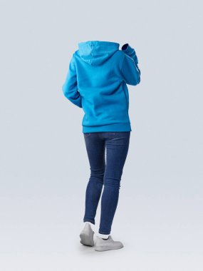 Parlak mavi kapüşonlu ve kot pantolonlu bir figür, hafif ışıklandırmalı, minimalist bir iç mekan ortamında günlük moda sergileyerek uzaklaşıyor..