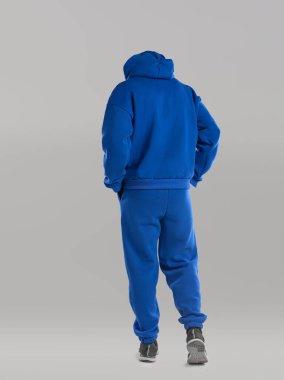 Arkası dönük, mavi kapüşonlu ve uyumlu eşofmanlı bir adam. Bu günlük kıyafet, basit gri bir arka plana göre ayarlanmış, modern sokak kıyafetleri sergileniyor..