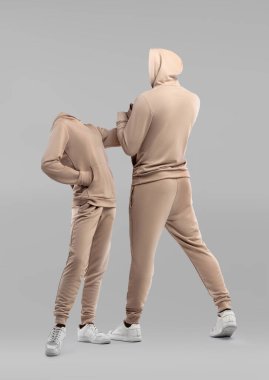 İki model minimalist bir stüdyoda bej kapüşonlu kıyafetler sergiliyor. Benzersiz sunum, modern tasarımı ve kıyafetlerin konforunu vurguluyor..