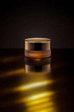 Altın bir kavanoz parlak bir yüzeyde, yumuşak bir yansıma yaratan sıcak ışıkla aydınlatılır. Koyu arkaplan zarif tasarımını geliştirir ve sakin bir ortam yaratır..