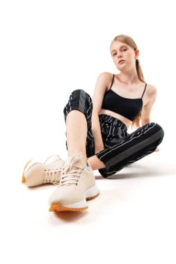 Genç bir kadın spor ayakkabılarıyla yerde otururken siyah bir bluz ve çizgili pantolon giyiyor. İyi aydınlatılmış bir stüdyo ortamında kendine güvenen bir poz veriyor..