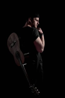 Bir müzisyen sırt üstü dinlenen bir gitarla güven içinde poz verir. Işıklandırma onun güçlü profilini vurguluyor, karanlığa karşı çarpıcı bir kontrast oluşturuyor..