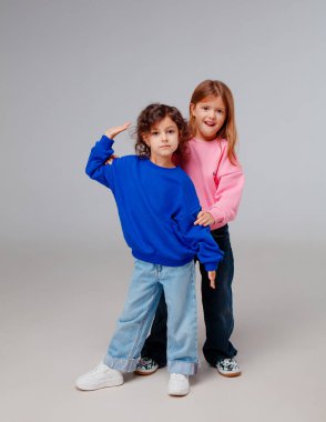 Renkli tişörtlerle yan yana duran iki kız, basit bir stüdyo arka planında neşeli bir arkadaşlık ve oyunculuk anı tasvir ediyorlar..