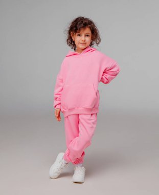 Bir çocuk bir eli kalçasında, canlı pembe bir eşofman ve beyaz bir spor ayakkabı giyiyor. Basit bir arkaplan karşısında neşeli bir ifade sergiliyor.