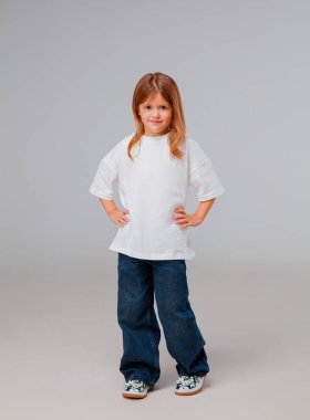 Bir çocuk, elleri kalçasında, beyaz bir gömlek ve kot pantolon giyerek güvenle ayakta duruyor. Arka plan, çocuğun neşeli ifadesini ve rahat duruşunu vurgulayan yumuşak bir gridir..