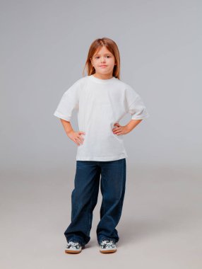 Genç bir çocuk, bol beyaz tişört ve kot pantolon giyerek kendinden emin bir şekilde ayakta duruyor. Tarafsız arka plan, deneğin neşeli ifadesini ve rahat duruşunu vurgular ve neşeli bir atmosfer yaratır..