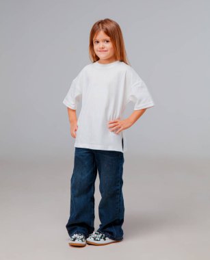 Bir çocuk gülümsüyor, bol beyaz gömlek ve kot pantolon giyiyor. Arka plan sade, kıyafetin rahat ve rahat stilini ve çocuğun neşeli ifadesini vurguluyor..